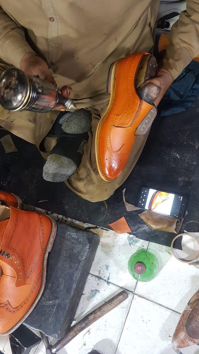 Op de afbeelding: Een schoenmaker werkt aan een paar oranje leren herenschoenen. De schoenen hebben decoratieve perforaties en een klassiek ontwerp. De schoenmaker gebruikt gereedschap om de schoen te vormen en te verfijnen, wat het vakmanschap laat zien.