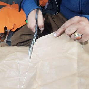 Op de afbeelding: Een persoon gebruikt een schaar om lichtbruin leer te snijden. Het leer heeft witte stiksels en wordt langs een gemarkeerde lijn gesneden. Oranje papieren stukken zijn op de achtergrond.