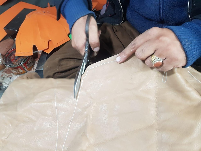 Op de afbeelding: Een persoon gebruikt een schaar om lichtbruin leer te snijden. Het leer heeft witte stiksels en wordt langs een gemarkeerde lijn gesneden. Oranje papieren stukken zijn op de achtergrond.
