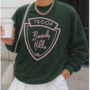May include: Dark green sweatshirt with the text "TROOP Beverly Hills" in pink, set within a shield design. The sweatshirt has long sleeves and a crew neck. The person is wearing a gold chain necklace and a white skirt.