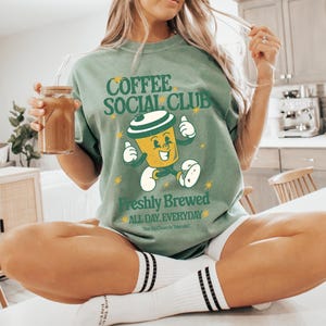 Könnte beinhalten: Salbeigrünes T-Shirt mit dem Text "Coffee Social Club" und einer Cartoon-Kaffeetasse. Das Shirt hat auch die Worte "Freshly Brewed All Day, Everyday". Die Person hält ein Glas mit einem Getränk.
