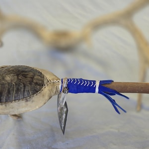 May include: A handcrafted Native American ceremonial rattle. The rattle features a turtle shell head, a wooden handle, and is adorned with blue leather accents and fringe. A pair of antlers are in the background.