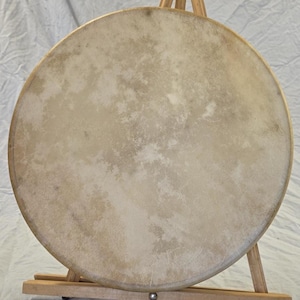 May include: A round, natural-toned drumhead with a light brown wooden frame. The drumhead is mounted on a wooden stand. The drumhead has a textured surface with variations in color.