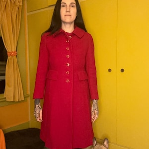 May include: A woman wearing a red coat with a collar and button closure. She is standing in front of a yellow wall with a dog sitting in front of her.