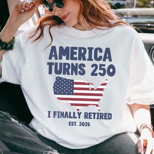 May include: White t-shirt with the text "AMERICA TURNS 250" in blue, and a red, white, and blue map of the United States. Below the map, the text reads "I FINALLY RETIRED EST. 2026".