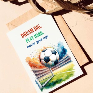 Puede incluir: Una ilustraci&oacute;n de acuarela de un bal&oacute;n de f&uacute;tbol volando hacia una porter&iacute;a. El fondo es una acuarela colorida de un estadio. El texto "DREAM BIG, PLAY HARD, never give up!" est&aacute; escrito en rojo, verde y azul.
