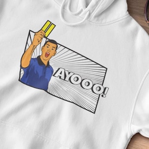 Puede incluir: Sudadera con capucha blanca con un gráfico de un árbitro mostrando una tarjeta amarilla y la palabra "AYOOO!". La imagen incluye zapatillas negras, un gorro verde y auriculares. La sudadera está sobre una superficie de madera.