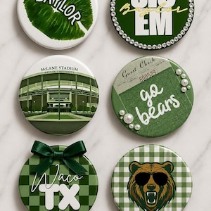 May include: Six circular buttons featuring Baylor University themes. Designs include a green leaf with 'BAYLOR', 'SIC EM' with rhinestones, McLane Stadium, a 'Guest Check' with 'go bears' and pearls, 'Waco TX' with a green bow, and a bear wearing sunglasses.