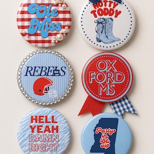 May include: Six circular pin-back buttons featuring various designs: a red and white gingham pattern with pearls, a white button with a navy bow and cowboy boots, a blue striped button with rhinestones, a red button with ribbons, a blue zebra print, and a blue Mississippi state shape.