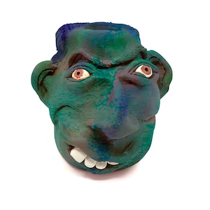 Puede incluir: Escultura de rostro de cerámica hecha a mano en tonos verdes y azules. La escultura presenta una amplia sonrisa, dientes visibles y rasgos faciales detallados, incluyendo ojos con iris marrones. Probablemente es un objeto decorativo o una obra de arte.