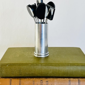 May include: A collection of black and white cocktail picks in a silver metal holder. The picks have geometric designs. The holder sits on top of two antique books with green and brown covers.
