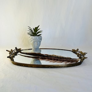 May include: An oval, antique-style mirror tray with a brass frame and decorative floral accents. A small white bust planter with a green succulent and a brown feather rest on the mirrored surface. The tray is on a white surface.