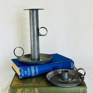May include: Three antique metal candle holders with ring handles and a fluted edge, resting on top of two vintage books. The books are blue and green, with gold lettering. The candle holders are a dark gray color.