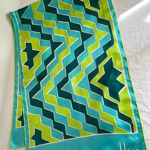 May include: A rectangular silk scarf with a geometric pattern in shades of teal, green, and white. The design features zigzags and star-like shapes. The scarf has a teal border and the signature "Usna" in the corner.