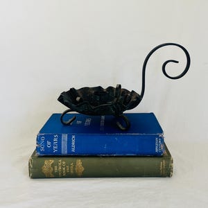 May include: A black metal candle holder with a scalloped edge, a central candle cup, and a curved handle, resting on two stacked hardcover books. The top book is blue with the text Song of Years, and the bottom book is green with the text The Victrola Book of the Opera.