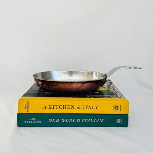 May include: A small copper-bottomed frying pan with a silver interior and handle rests on two books. The books are titled "A Kitchen in Italy" and "Old World Italian". The background is a plain white surface.