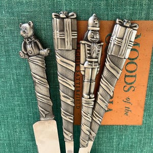 May include: Silver serving utensils with decorative handles. The handles feature a teddy bear, gift boxes, and a nutcracker design. The utensils are displayed on a green surface with a brown tag that has text.