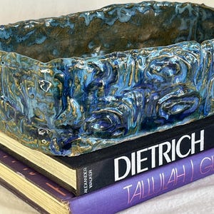 May include: A rectangular ceramic planter with a textured, blue and gold glaze. The planter sits atop two books with the titles "DIETRICH" and "TALLULAH!" visible. The planter's surface has a rough, organic appearance.