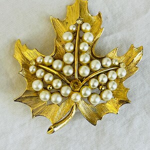 May include: Gold-toned leaf-shaped brooch with a textured surface. The brooch is adorned with faux pearls and a small, clear rhinestone. The leaf design has detailed veins and a stem.