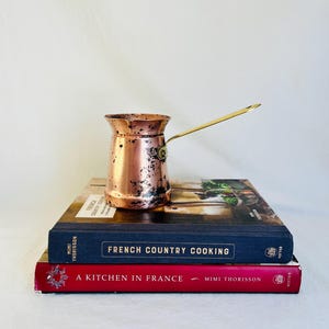May include: A copper coffee pot with a long brass handle rests atop a stack of two cookbooks. The books are titled "French Country Cooking" and "A Kitchen in France". The coffee pot has a tarnished finish.