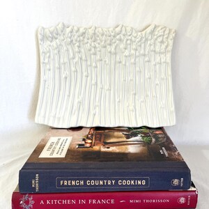 May include: A rectangular white ceramic asparagus serving dish with a textured, raised relief pattern of asparagus stalks. The dish is resting on two stacked hardcover books titled French Country Cooking and A Kitchen in France by Mimi Thorisson.