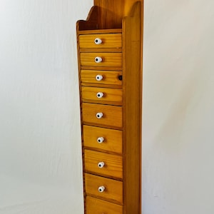 May include: A tall, narrow wooden storage unit featuring nine stacked drawers. Each drawer is equipped with a small, round white knob with a black center. The unit has a light brown wood grain finish and a curved, arched top piece with a hole for hanging.