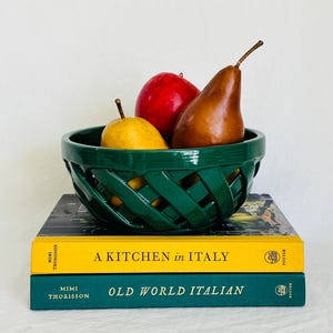 May include: A dark green ceramic bowl with a woven lattice design containing a yellow pear, a red apple, and a brown pear. The bowl rests on two stacked books titled A Kitchen in Italy and Old World Italian by Mimi Thorisson.