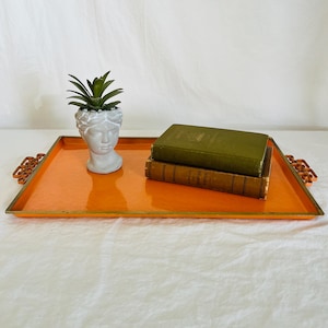 May include: A rectangular orange tray with a metallic gold rim and ornate handles. On the tray sits a white ceramic head-shaped planter containing a green succulent and a stack of two olive green hardcover books with gold lettering.