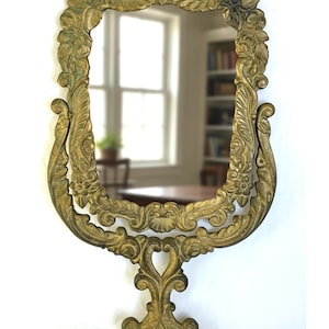 May include: Ornate gold-colored vanity mirror with an oval-shaped frame. The frame features intricate floral and scrollwork designs. The mirror is supported by a decorative base with a heart-shaped detail. The mirror reflects a room with a window and bookshelves.
