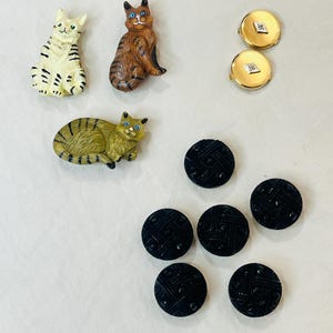 May include: A collection of sewing notions including three cat-shaped buttons in cream, brown, and olive green, two round gold-tone buttons with a central diamond-shaped accent, and five round black buttons featuring a textured cross-hatch pattern.