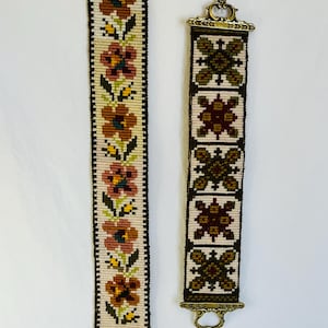 May include: Two decorative textile wall hangings. The left one features a floral pattern with brown, red, and yellow flowers on a cream background. The right one has a geometric design with similar colors. Both have metal hardware.