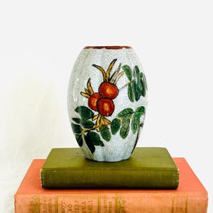 May include: A white ceramic vase with a crackled glaze and a hand-painted design of red berries and green leaves. The vase is sitting on top of two books.