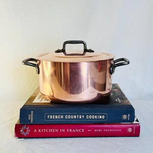 May include: A copper cooking pot with a lid and black handles sits atop a stack of two cookbooks. The cookbooks are titled "French Country Cooking" and "A Kitchen in France". The pot has a shiny, reflective surface.
