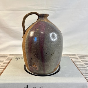 May include: A small, brown, gray, and purple ceramic pitcher with a handle. The pitcher has a textured surface and a slightly rounded shape.