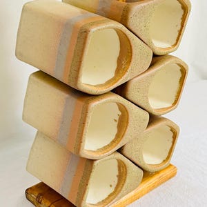 May include: A ceramic wine rack featuring six rectangular, cream-colored holders with soft peach and grey horizontal bands. The holders are arranged in a stacked, staggered formation and are mounted on a rectangular wooden base.