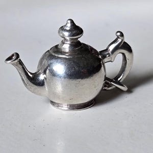 May include: A silver teapot with a rounded body and a curved spout. The teapot has a decorative lid with a knob on top.