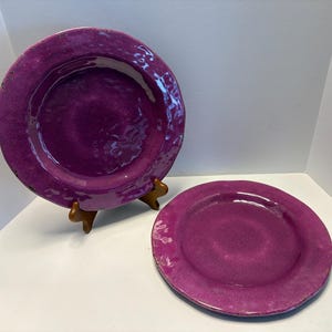 May include: Two purple ceramic plates. One plate is larger and propped up on a wooden stand. The plates have a textured surface and a glossy finish. The colour is a deep, rich shade of purple.