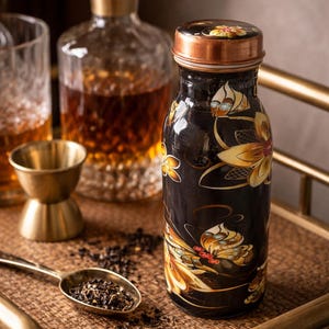 May include: A black decorative bottle with a copper lid and floral design. The bottle is on a woven tray with a gold spoon and a jigger. A glass decanter and a glass are in the background.