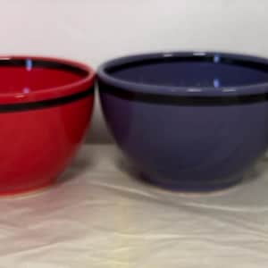 May include: A set of four ceramic bowls arranged in a row. Two bowls are lime green with a black scalloped pattern and a black rim. One bowl is solid red with a black rim. One bowl is solid purple with a black rim. All bowls have a smooth finish.