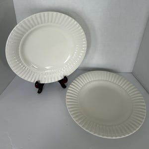 May include: Two off-white ceramic plates with a ribbed design around the edges. One plate is on a dark brown stand. The plates have a simple, elegant design, suitable for serving food. The plates are round and oval.