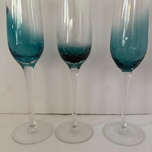 Peut inclure: Trois flûtes à champagne transparentes avec un effet ombré turquoise. La base de chaque verre présente un motif craquelé. Les tiges sont longues et fines, et les bases sont rondes. Les verres sont vides et posés sur un fond blanc.