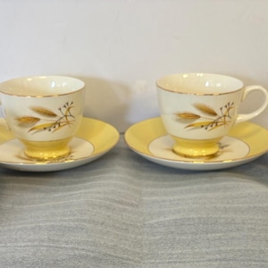 Puede incluir: Cuatro tazas y platillos vintage. Las tazas son de color crema con ribetes dorados y un diseño similar al trigo. Los platillos son amarillos con un centro color crema. Las tazas se asientan sobre una base amarilla.