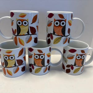 May include: Five white ceramic mugs with owl and leaf designs. Each mug features a different owl illustration in orange, yellow, and black, perched on a branch with autumn leaves. The mugs have large white handles.