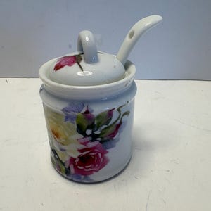 May include: A white ceramic sugar bowl with a lid and spoon. The bowl and lid are decorated with a floral pattern of pink, yellow, and purple roses. The spoon rests on the lid, ready for use.