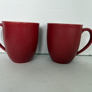 Peut inclure: Deux tasses en céramique rouge avec des anses incurvées. Les tasses ont une finition lisse et mate et une forme simple et arrondie. L'intérieur des tasses semble être d'une nuance de rouge plus claire. Ces tasses conviennent au café, au thé ou à d'autres boissons.