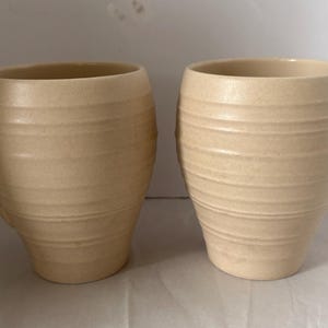 May include: Two matching mugs with a light beige, speckled finish. Each mug features a rounded, barrel-like body with horizontal ridges encircling the exterior and a curved handle on the side. The surface has a matte, textured appearance.
