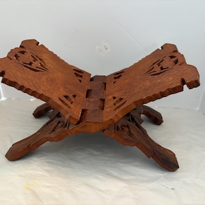 May include: A wooden book stand with a dark brown finish. The stand is open, with carved floral designs on the top edges. The legs are also carved and provide support. The stand is designed to hold a book open for reading.