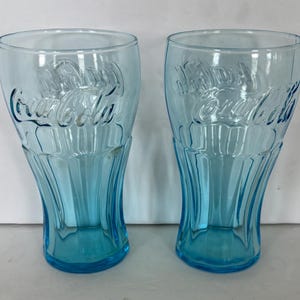 May include: Two light blue Coca-Cola glasses. The glasses have the Coca-Cola logo embossed on the front. The glasses have a classic shape with a flared base and a curved body. The glasses are empty and sitting on a white surface.