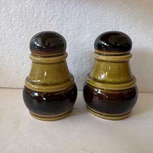 May include: A pair of ceramic salt and pepper shakers. The shakers are brown, olive green, and tan. The top of each shaker is a dark brown, with a series of holes. The middle section is olive green, and the bottom is dark brown.