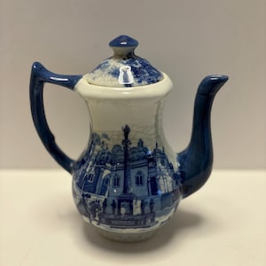 Puede incluir: Una tetera de cerámica azul y blanca con tapa. La tetera presenta una detallada escena de paisaje azul alrededor del cuerpo y en la tapa. El asa y el pico son de color azul liso. La tetera es de estilo vintage.